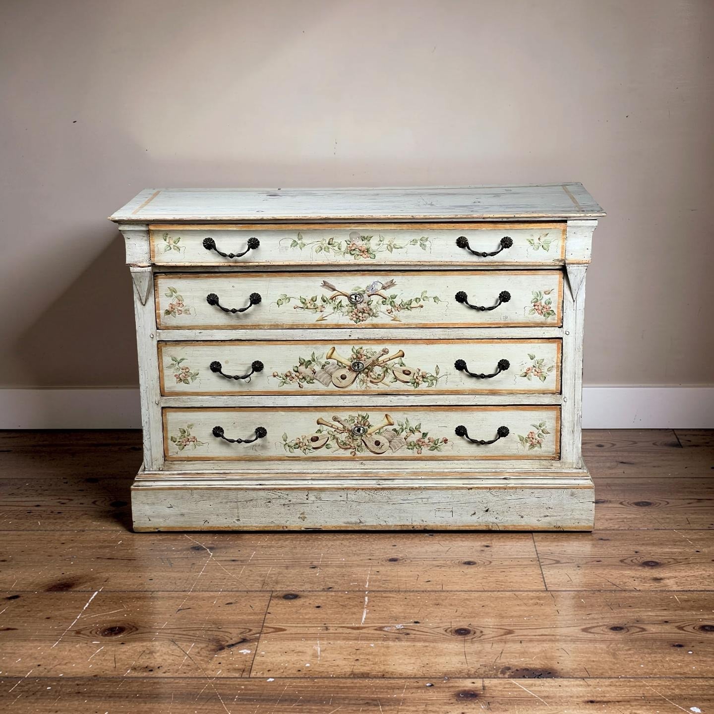 An Antique Italian Paint Decorated Four Drawer Commode, c. Early 19th Century, with floral and scrollwork designs in a distressed pale blue finish, stands on a wooden floor against a plain beige wall. Brand: None/Unknown.