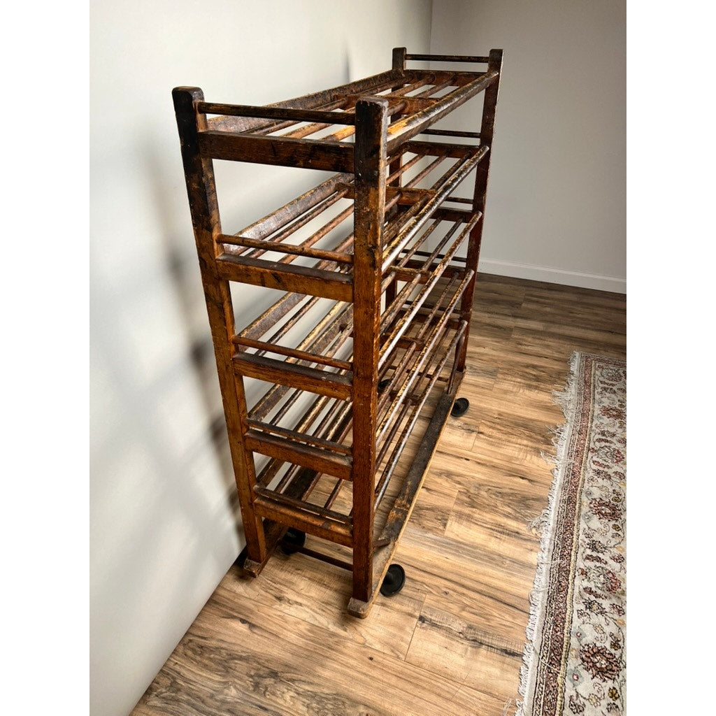 An Antique Oak Shoe Factory Rolling Rack on original industrial casters (c. late 19th century) stands on a wooden floor near a white wall and patterned rug, featuring multiple shelves and a worn, rustic finish. Brand unknown.