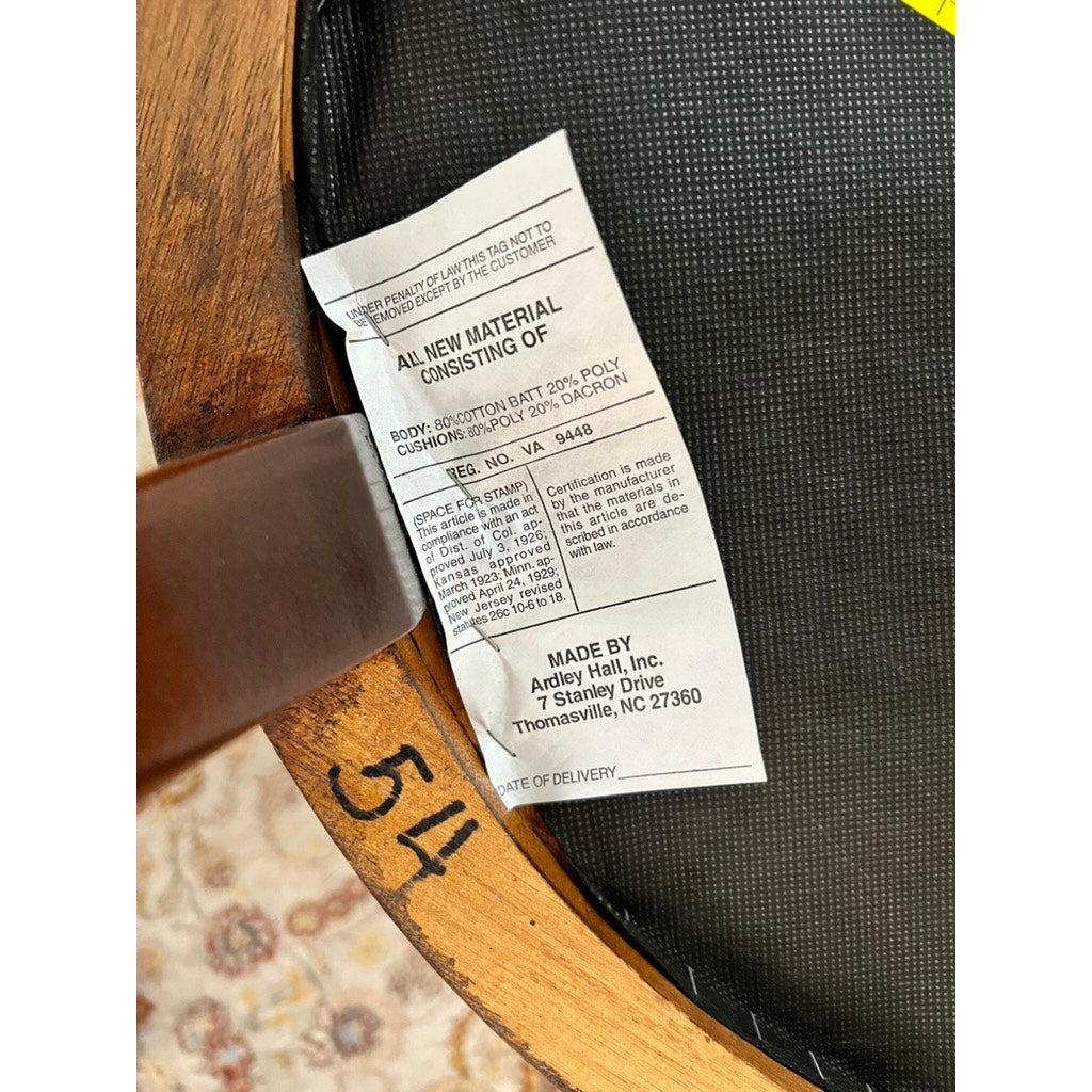 A close-up of a tag under an Ardley Hall Vintage Pair Wheel Back Side Chair shows materials and company info. The chair’s classic English design, wood frame, and patterned carpet are also visible.