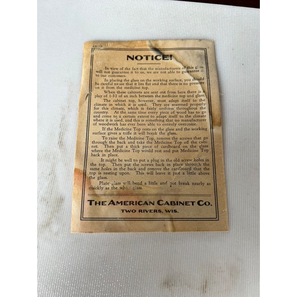 A vintage, stained instruction card from The American Cabinet Co., makers of the Antique Mahogany Multi-drawer Dental Cabinet c. Early 20th Century, offers care directions. The card is displayed on a white textured surface.