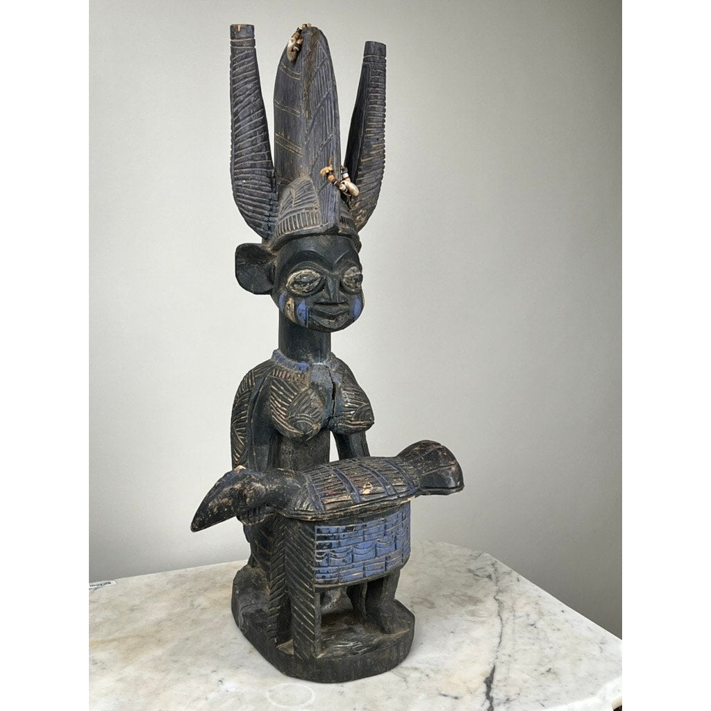 An Antique Igbo Ikenga Shrine Figure, likely from Nigeria, carved from wood and featuring a tall headdress, is displayed holding an animal-shaped object on a marble surface against a plain background. Brand: Unknown.