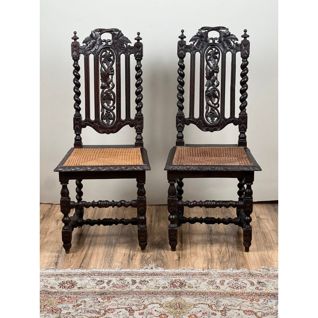 A pair of antique French walnut hunting chairs from the late 19th century, featuring intricately carved backs and twisted legs, are side by side on a wooden floor with part of a patterned rug visible below.