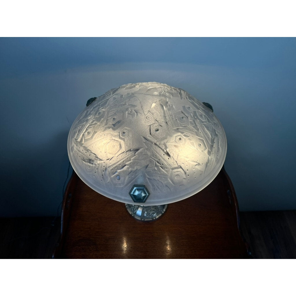 A Vintage French Art Deco Frosted Glass Table Lamp by Muller Frères Lunéville c. 1920 sits on a wooden surface, its geometric frosted glass shade glowing softly and casting teal accents against a blue-gray wall.