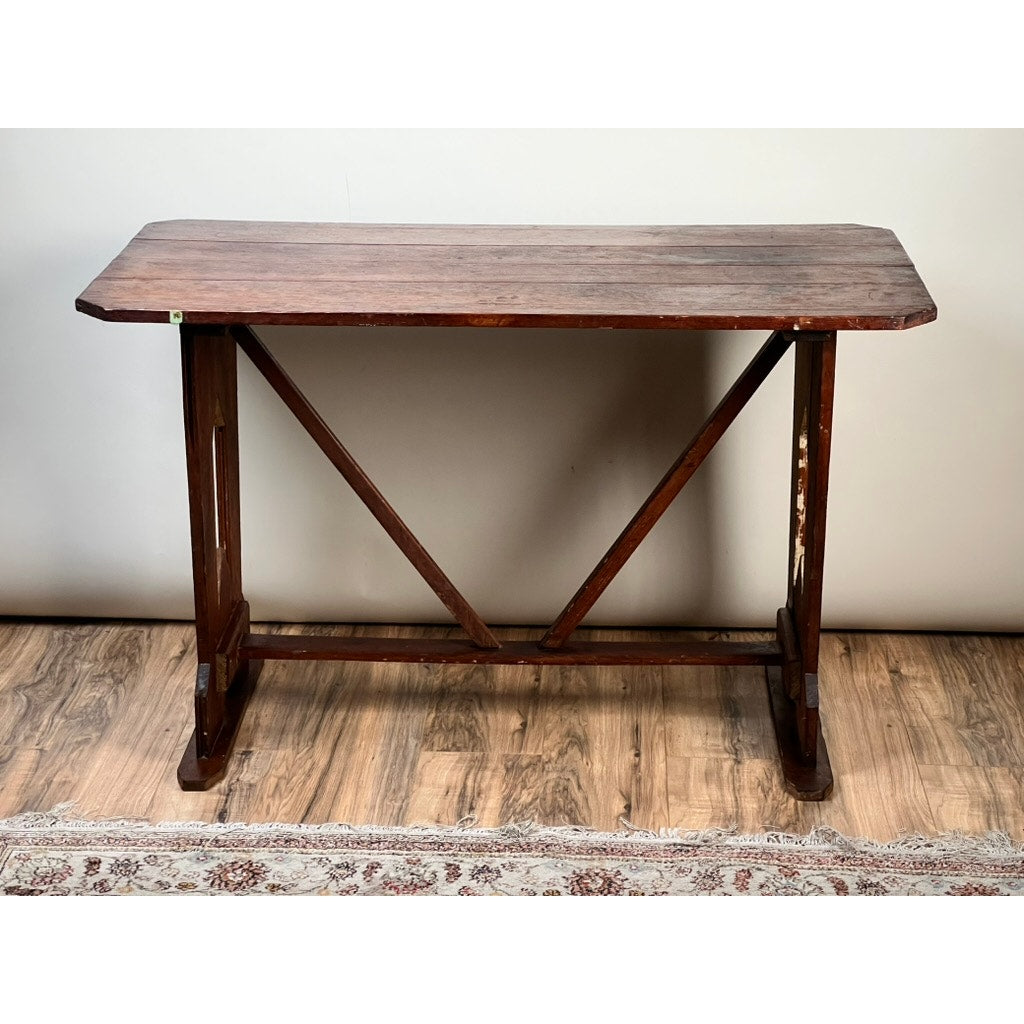 The Antique Rustic Italian Fratino Trestle Table (c. late 19th century) has a rectangular solid wood top, angled edges, and sturdy legs with diagonal supports, set on a wooden floor beside a patterned rug.