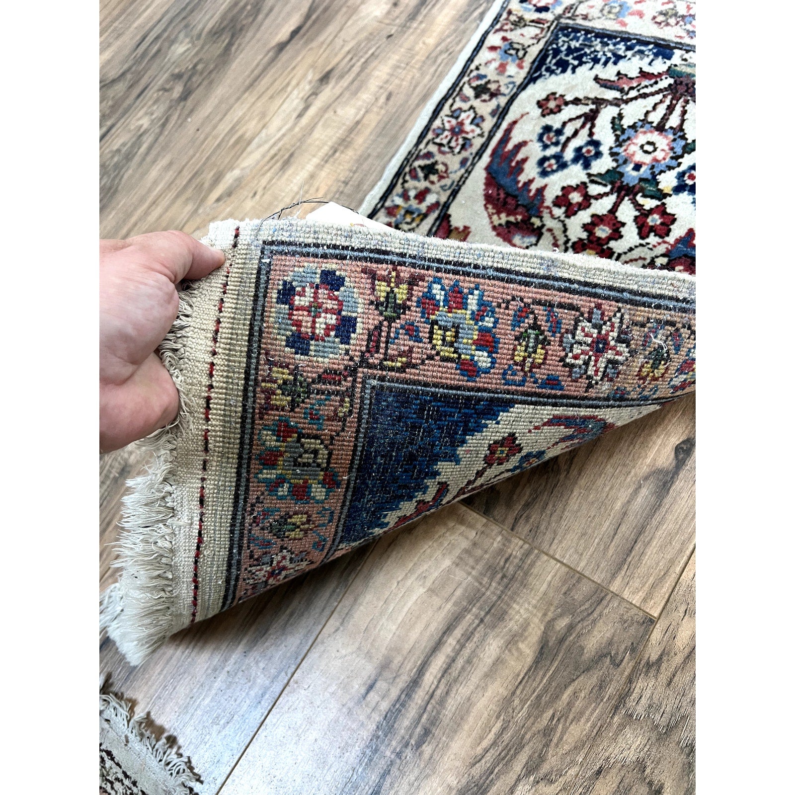 A hand lifts the corner of a Vintage Pair of Hand-Knotted Oriental Tree of Life Scatter Rug (1.5 x 2.5), revealing intricate blue, red, and beige floral details on its underside atop a wooden floor. Brand: None/Unknown.