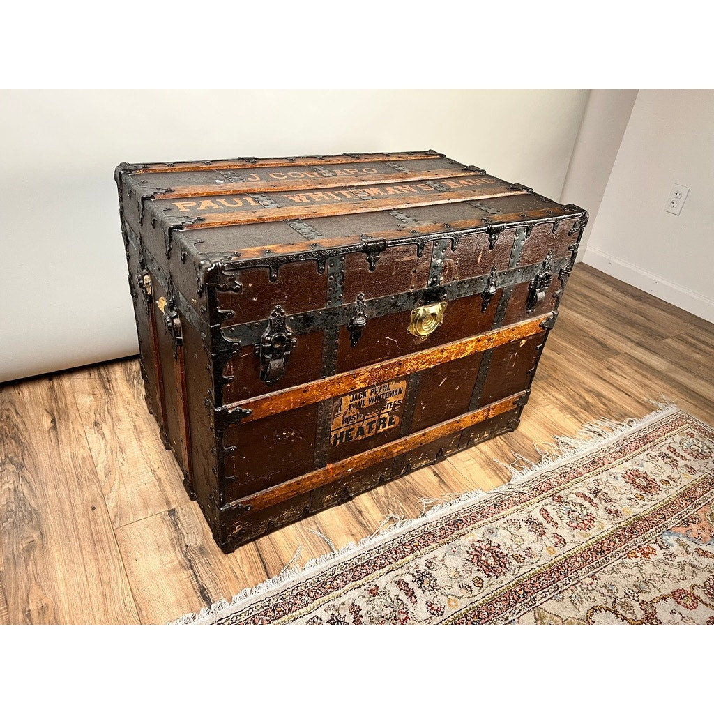 A vintage CA Taylor Touring Trunk of John B. Cordaro, Paul Whiteman Orchestra reed player (c. 1930s), with black metal accents and latches, rests on a wooden floor beside a patterned rug and against a white wall.