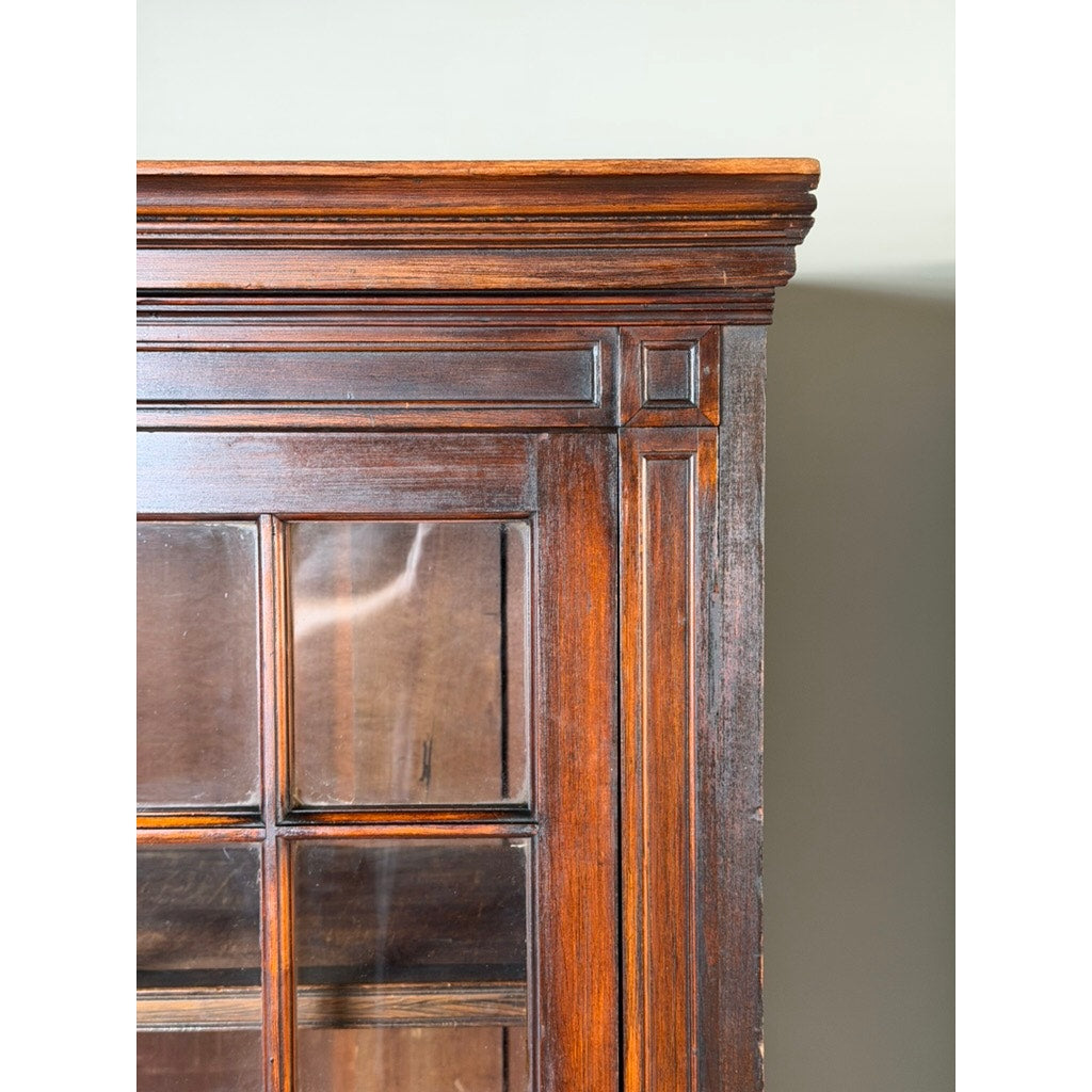 Close-up of the top right corner of a Pair of Mid-19th Century New England Cherry Glazed Display Cupboards by Pearl City Antiques, c. 1840–1860, featuring dark wooden construction with glass panels and decorative trim, reminiscent of early American cupboards, set against a light gray wall.