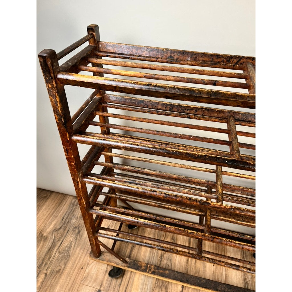 An Antique Oak Shoe Factory Rolling Rack on original industrial casters from the late 19th century, featuring aged wood and multiple slats, stands on a wooden floor against a plain light-colored wall.