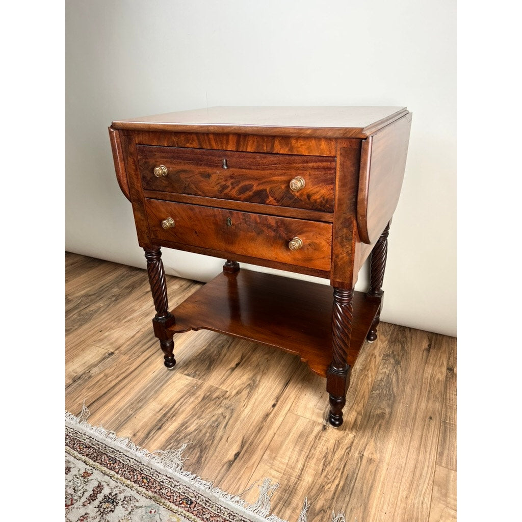An Antique Federal Mahogany Two Drawer Work Stand with Lions Head Pulls c. 1820 stands on a wooden floor beside a patterned rug, featuring drop-leaf sides and ornate twisted legs.