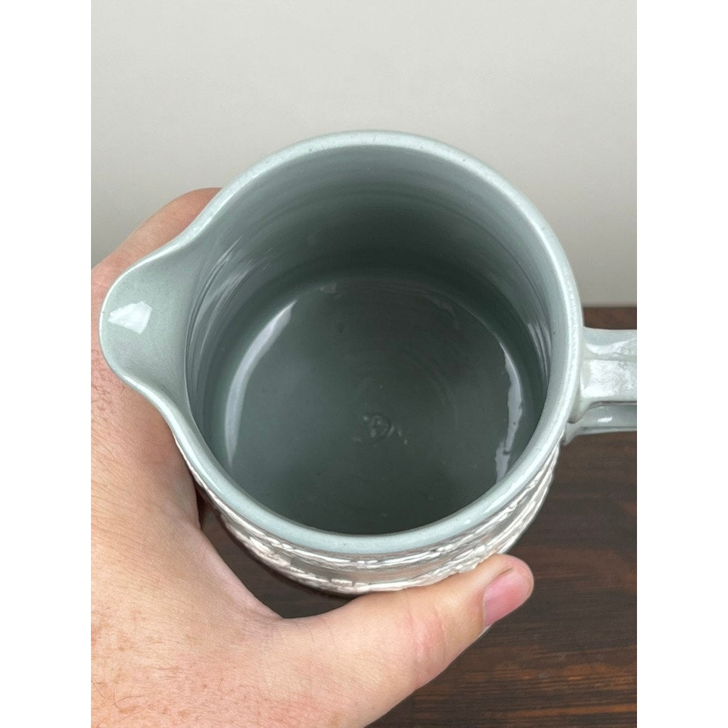 A hand holds a Vintage Copeland Spode English Jasperware Fox Hunt Pitcher, light gray with a handle, seen from above as it rests empty on a dark wooden surface.