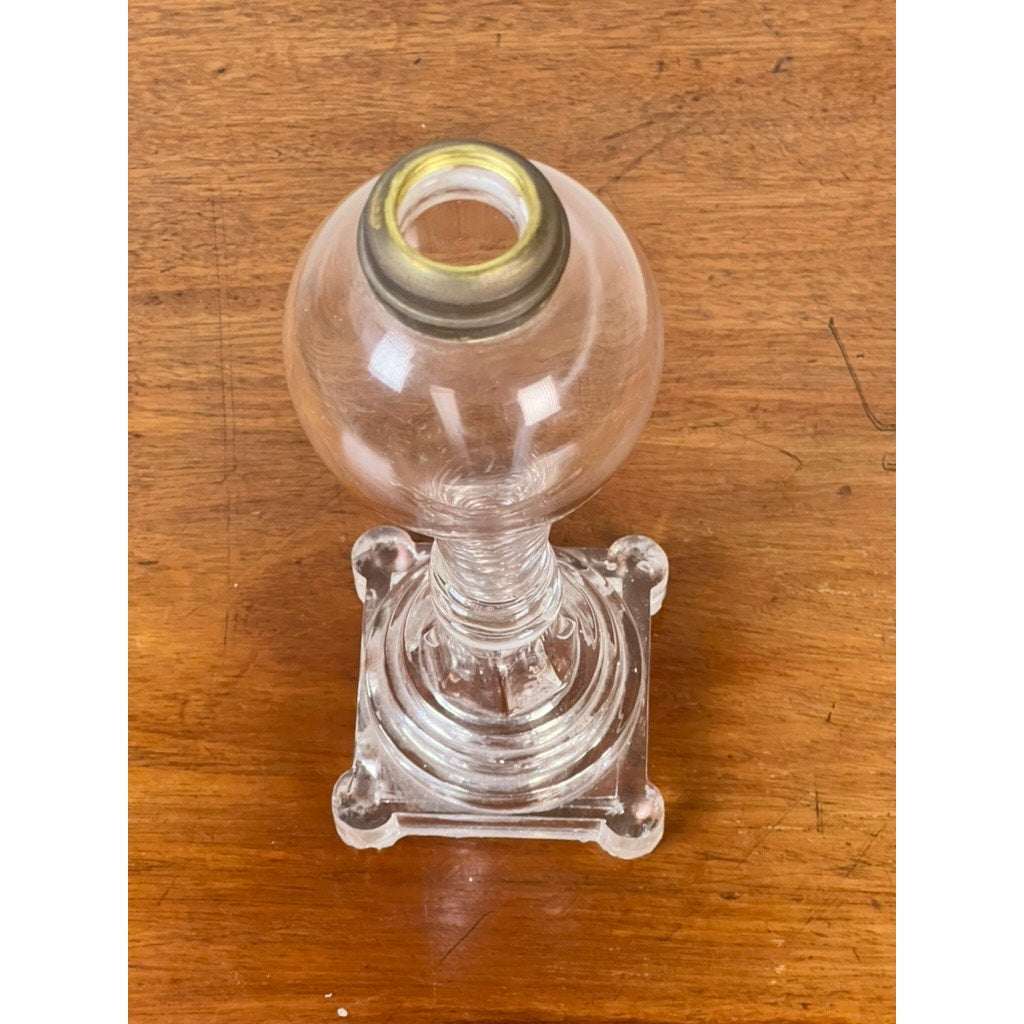 An Attributed Boston & Sandwich pressed glass boudoir fluid lamp, circa 1850—featuring a clear round reservoir, metal collar, and square base—is displayed on a wooden surface, exemplifying early American lighting design.