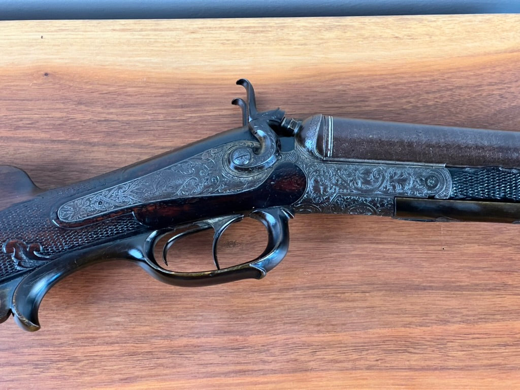Close-up of the Antique Christoph Funke Suhl German Damascus Double Barrel Hammer Shotgun, c. 1880, from Pearl City Antiques—showcasing ornate engravings on the metal receiver and trigger guard, striking Damascus barrels, and a carved stock, displayed on a wooden surface.