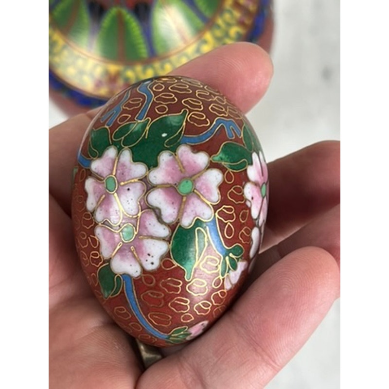 A hand displays a Vintage Chinese Cloisonné Egg with a red background, pink flowers, green leaves, and gold accents. The enamel-style decorative piece is shown with a colorful, blurred object in the background. Brand: Unknown.