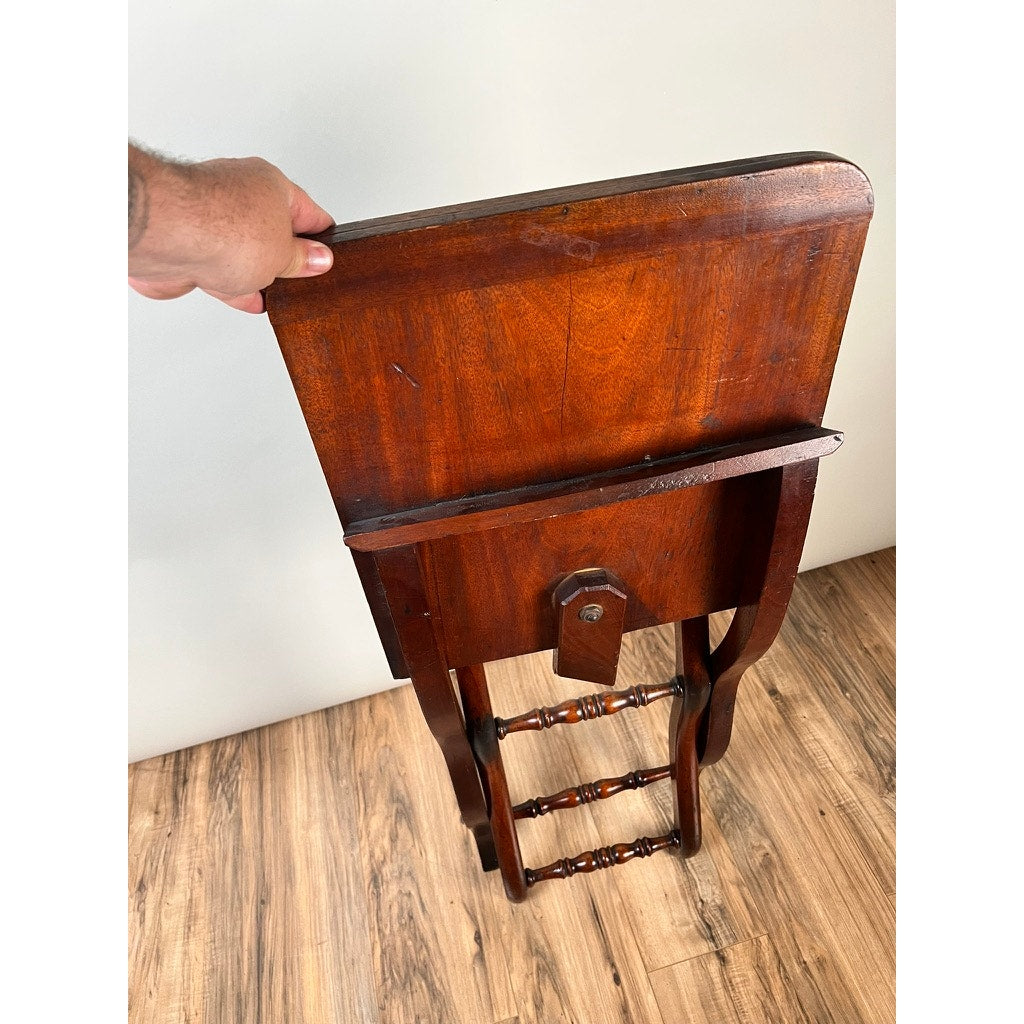 A person holds an Antique English Regency Mahogany Folding Coaching or Sporting Table (c. 1825–1845) by the top, showing its back and folded legs, on a light wood floor against a plain white wall. Brand: Unknown.