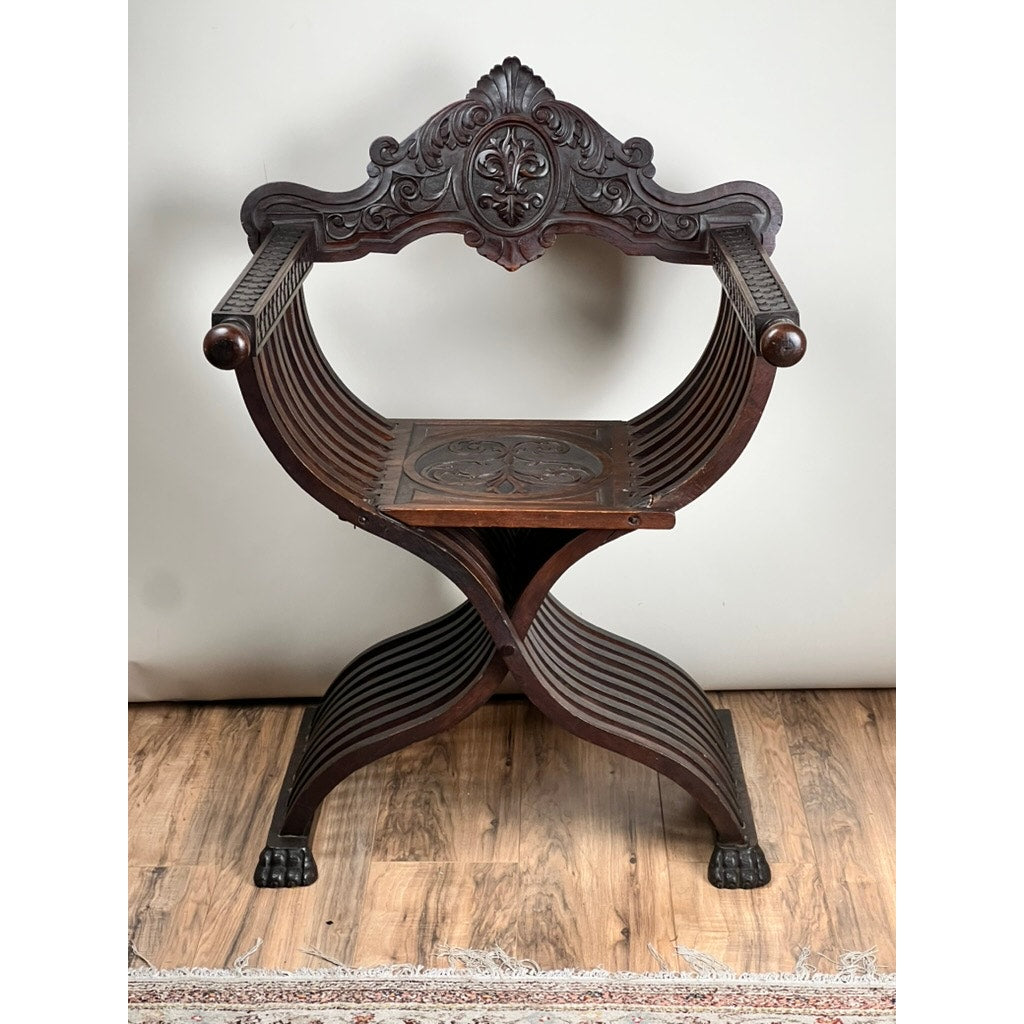 An Antique Italian Renaissance Walnut Savonarola Chair (late 19th c.) with an intricately carved backrest, curved X-frame, and lion paw feet sits on a wooden floor near a rug.