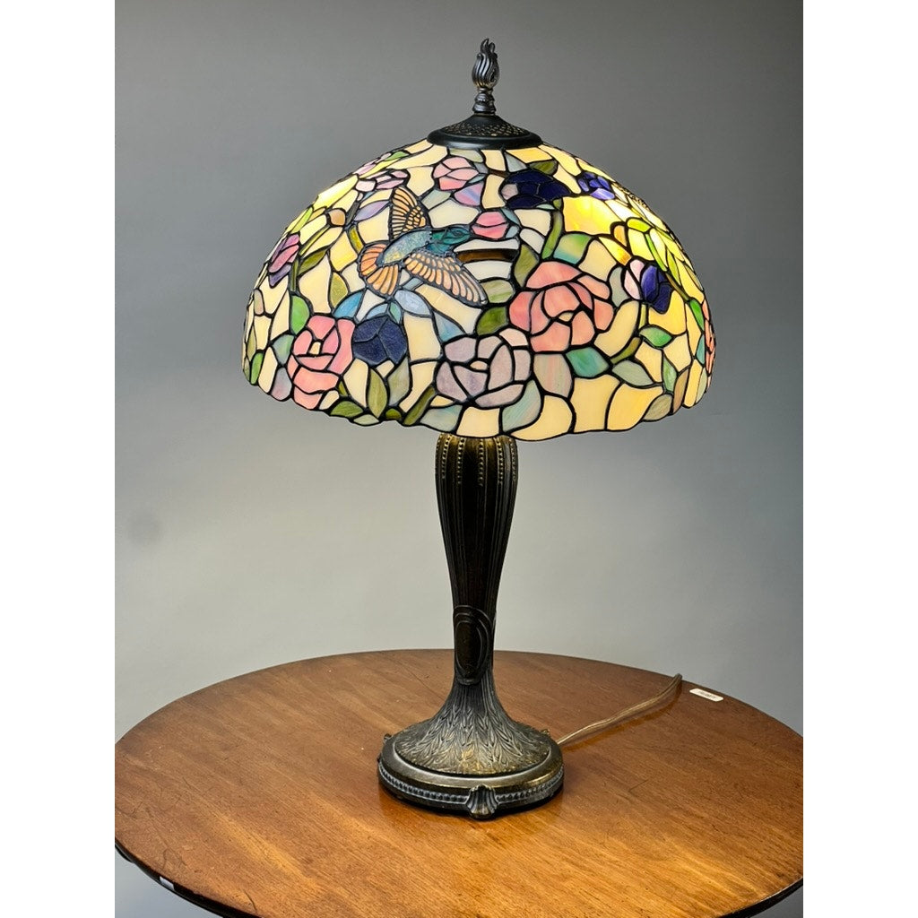 A Vintage Tiffany Style Floral Stained Glass Table Lamp with a colorful floral and hummingbird shade and a dark ornate base sits on a round wooden table against a plain gray background. Brand: None/Unknown.