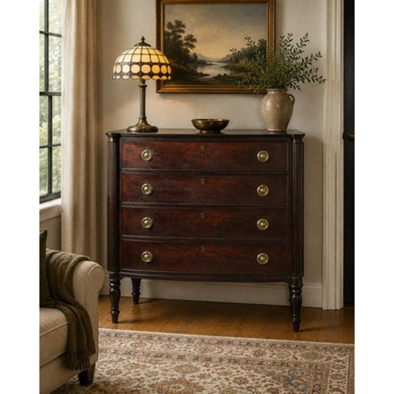 A vintage vignette features an Early 19th Century New England Federal Mahogany Bow-Front Chest of Drawers, c. 1810–1825, styled with four drawers and complemented by a brass lamp with a stained-glass shade, a decorative bowl, a vase filled with greenery, and a landscape painting on the wall. The arrangement is situated beside a window and an armchair atop a patterned rug.
