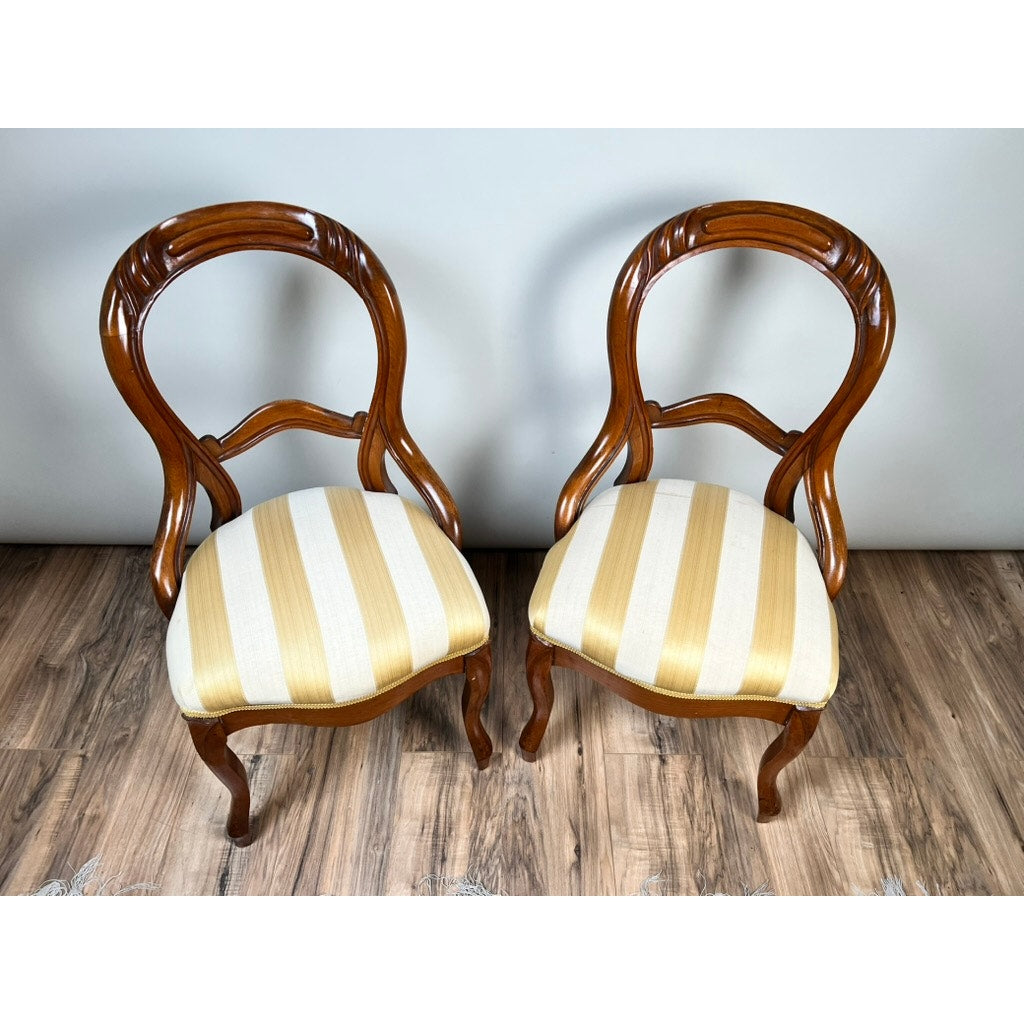 A pair of antique Victorian balloon back chairs from the late 19th century, both featuring carved details and cream and gold striped upholstered seats, are set on a wooden floor against a plain light-colored wall.