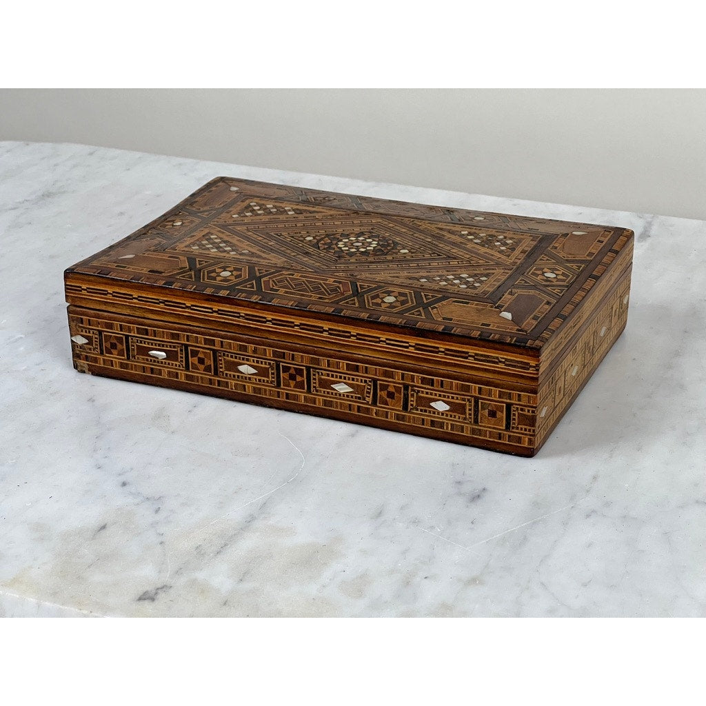 An Antique Persian (likely Syrian) Khatam wood inlaid jewelry box from the early 20th century, featuring intricate brown and beige geometric patterns, rests elegantly on a white marble surface. Brand unknown.