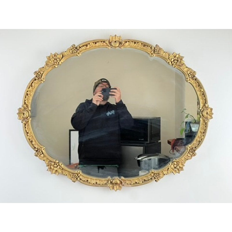 A person in dark clothing takes a selfie in an Antique Gilt Wood Oval Mirror c. Early 20th Century, mounted on a white wall, with kitchen appliances and a small plant visible in the reflection.