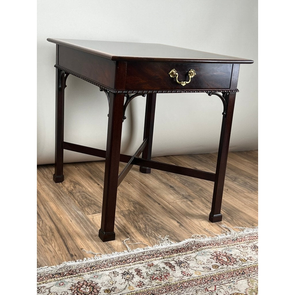 The Stickley Vintage Federal Style X Stretcher One Drawer End Table, featuring dark wood, a brass-handled drawer, decorative trim, and straight legs, stands on a wood floor beside a patterned rug against a light wall.
