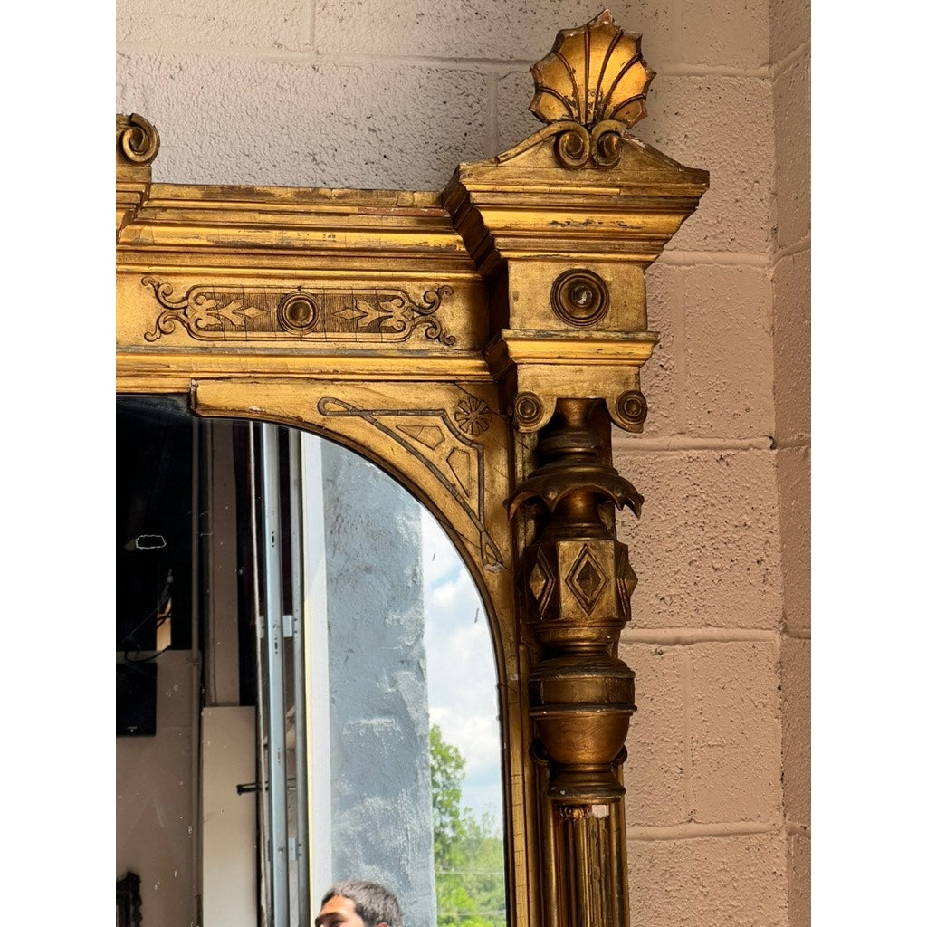 Exceptional Monumental Victorian Renaissance Revival Pier Mirror, Circa 1870–1885, featuring an ornate gold giltwood frame with detailed carvings and a shell motif at the upper corner, set against a textured beige brick wall. The mirror subtly reflects part of a window and the head of a person.