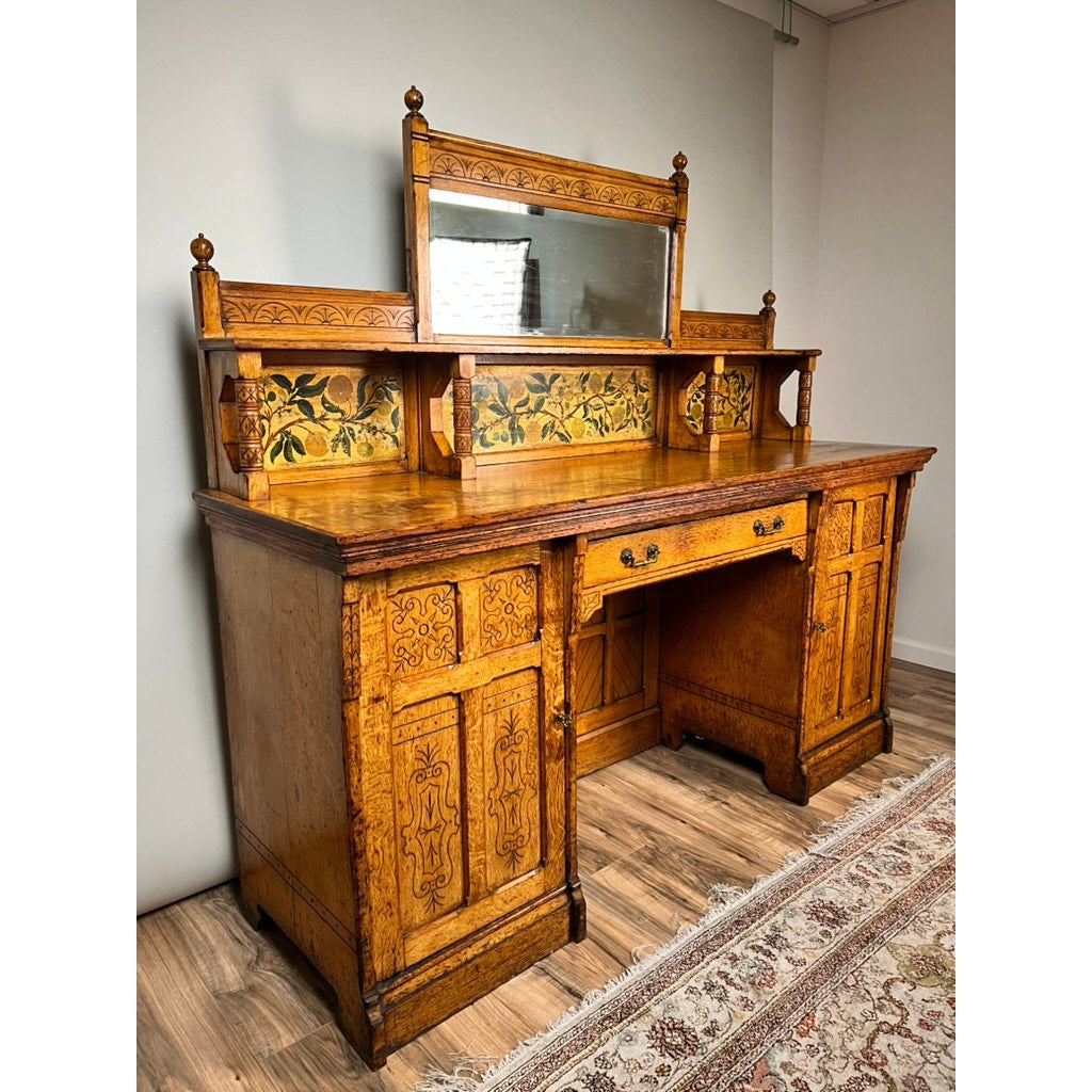 Antique Victorian Aesthetic Movement oak desk in original late 19th-century finish, featuring intricate carvings, floral painted panels, and a decorative mirror, displayed against a plain wall on a wooden floor with part of a patterned rug visible.