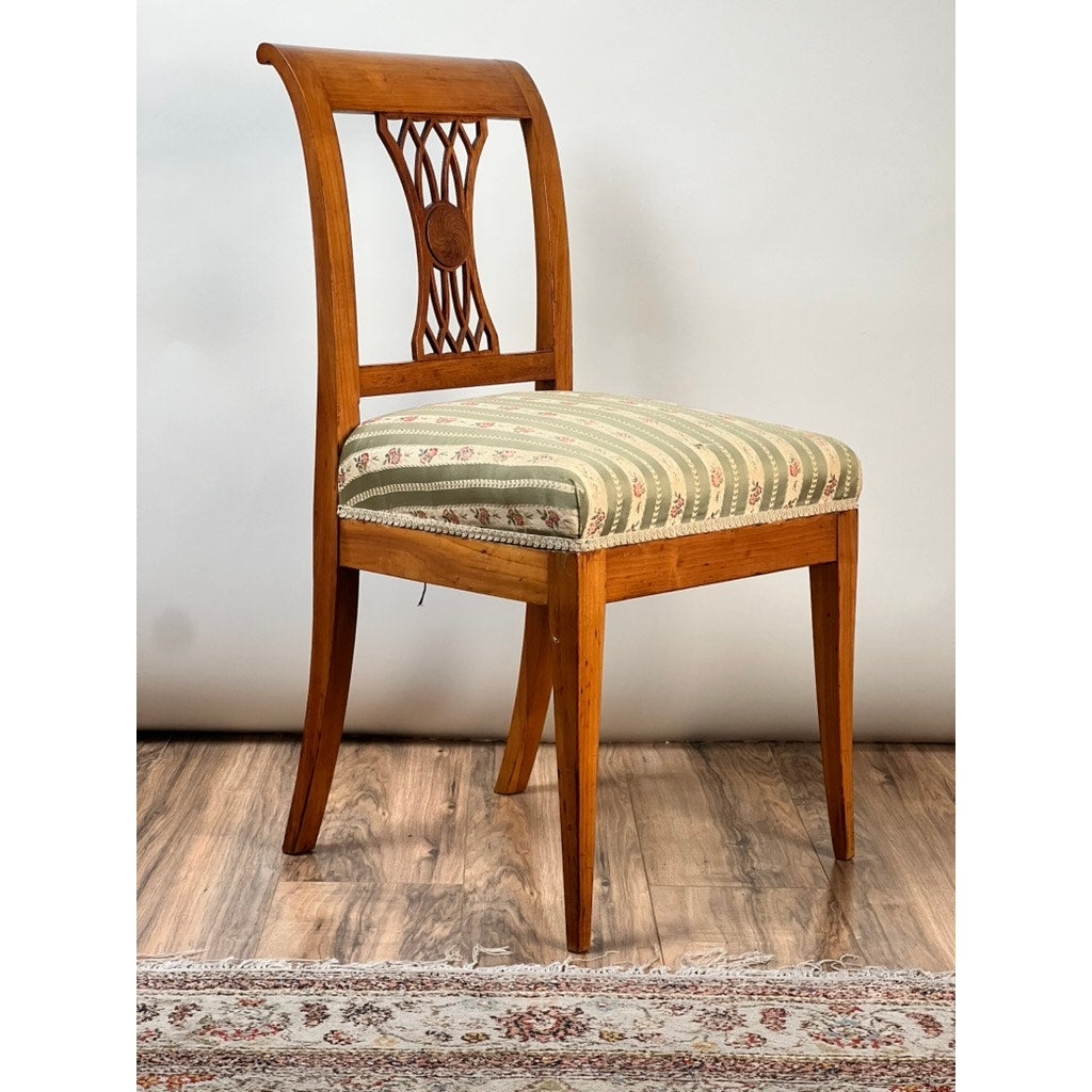 An Antique German Biedermeier Style Cherry Side Chair (early 20th c.) with a carved backrest and green-cream striped floral padded seat stands on a wood floor near a patterned rug. Brand: None/Unknown.