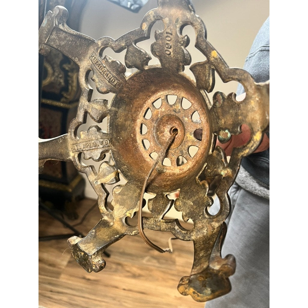 A close-up of the cast iron base of an Antique Art Deco Floor Lamp by Rembrandt, with ornate cutout patterns, visible patina, and a protruding wire. Part of a hand and a wooden floor are also shown.