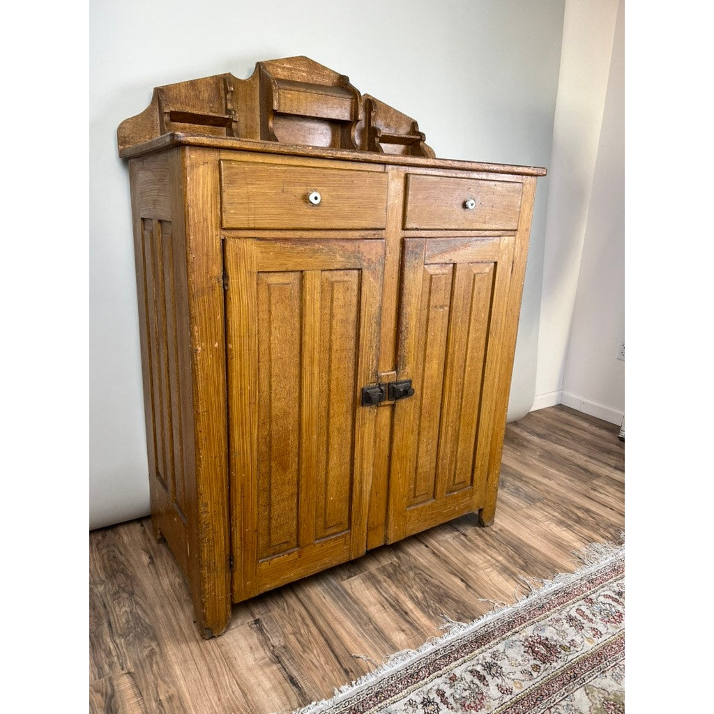 An Antique Victorian Country Grain Painted Jelly Cupboard c. 1870, with two drawers, paneled doors, metal handles, and a decorative top shelf, sits on a wooden floor beside a patterned rug. Brand: None/Unknown.