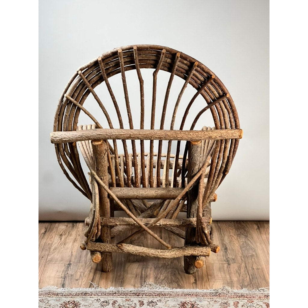 The Vintage Adirondack Bent Willow Arm Chair features a circular twig seat and rounded backrest made from natural, unpolished branches; it rests on a wooden floor with a patterned rug partially visible below. Brand: None/Unknown.