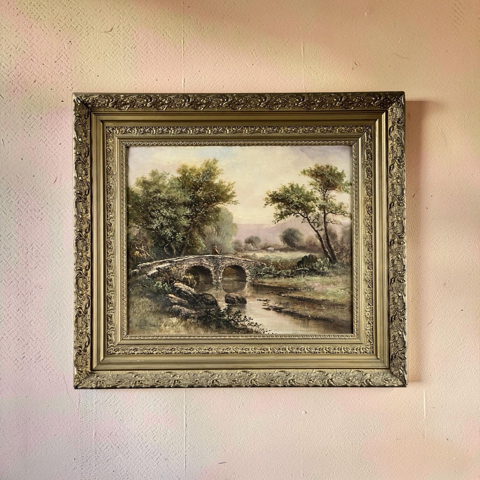 An antique early 20th century oil on canvas painting, “Spectacle Bridge,” signed Arthur Davis lower left, depicts a stone bridge over a stream in lush countryside and is displayed on a softly textured, light-colored wall.