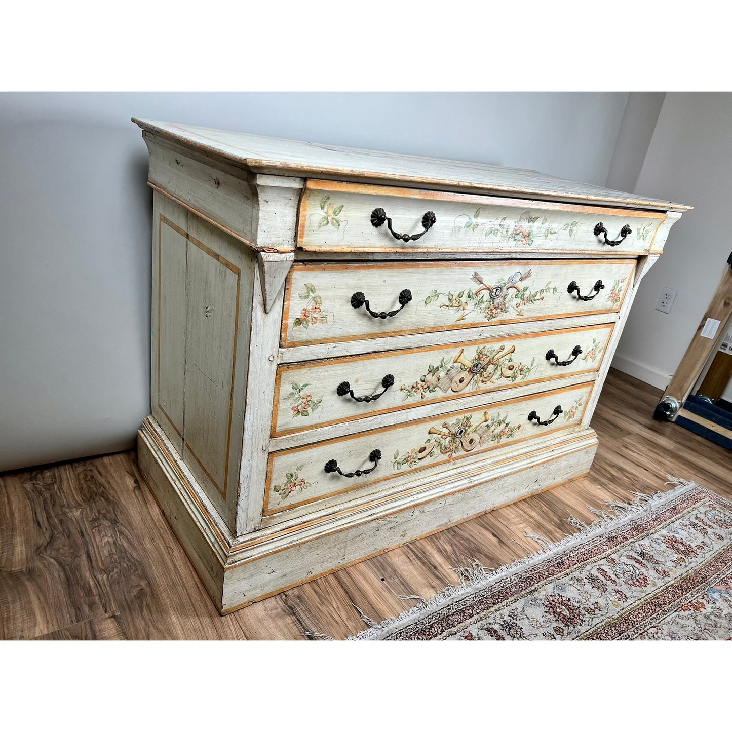 An Antique Italian paint-decorated four-drawer commode (early 19th century) with floral hand-painted designs and black metal handles stands on a wooden floor near a patterned rug in a bright room. Brand: Unknown.