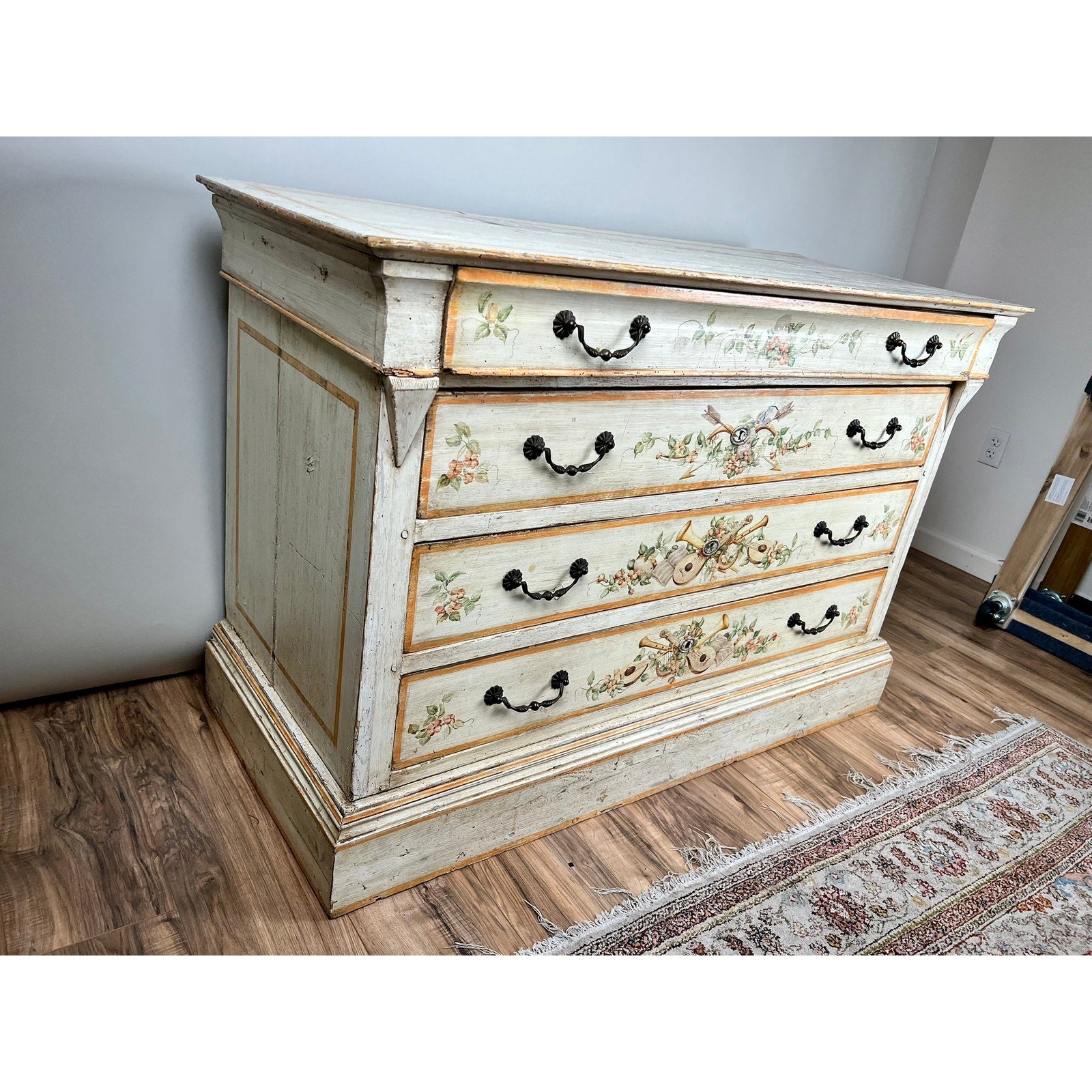 An Antique Italian paint-decorated four-drawer commode (early 19th century) with floral hand-painted designs and black metal handles stands on a wooden floor near a patterned rug in a bright room. Brand: Unknown.