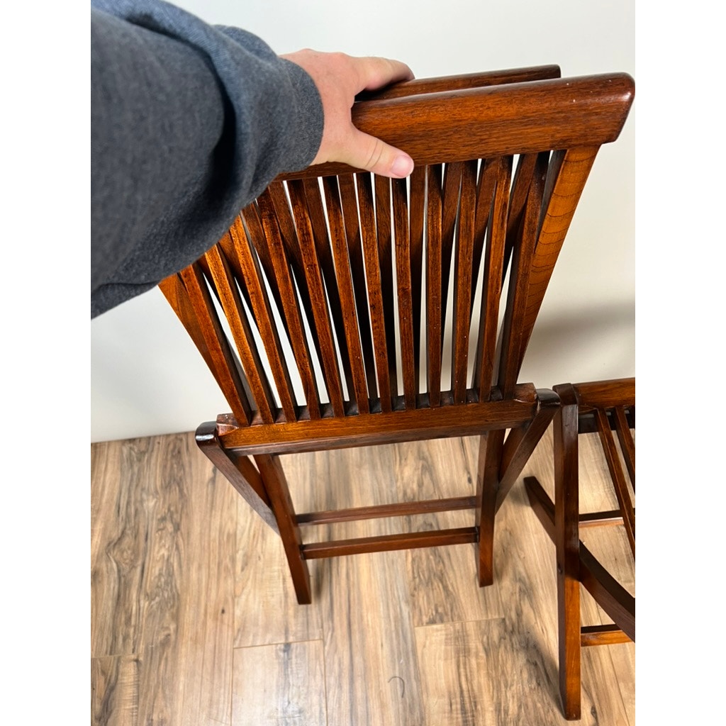 A person in a gray sleeve holds the top of a Pair Vintage Mid-Century Modern Style Teak Folding Chairs, seen from above on a wooden floor, with the second matching folding chair partially visible to the right. Brand: None/Unknown.