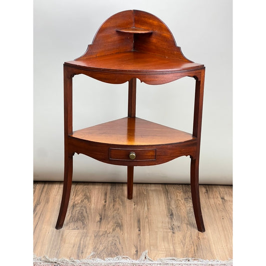 An Antique George III Mahogany Corner Wash Stand from the early 19th century, featuring a curved top shelf, lower shelf, small central drawer with round knob, and four curved legs, is displayed on a wooden floor against a neutral background.