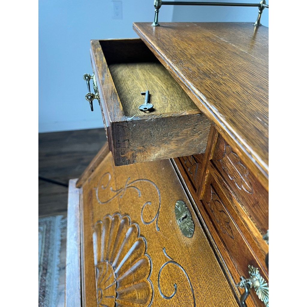 An open drawer revealing a vintage metal key inside an Antique Victorian Oak Ladies Writing Desk c. 1900, adorned with ornate carvings and brass hardware, stands on a wooden floor near a patterned rug.