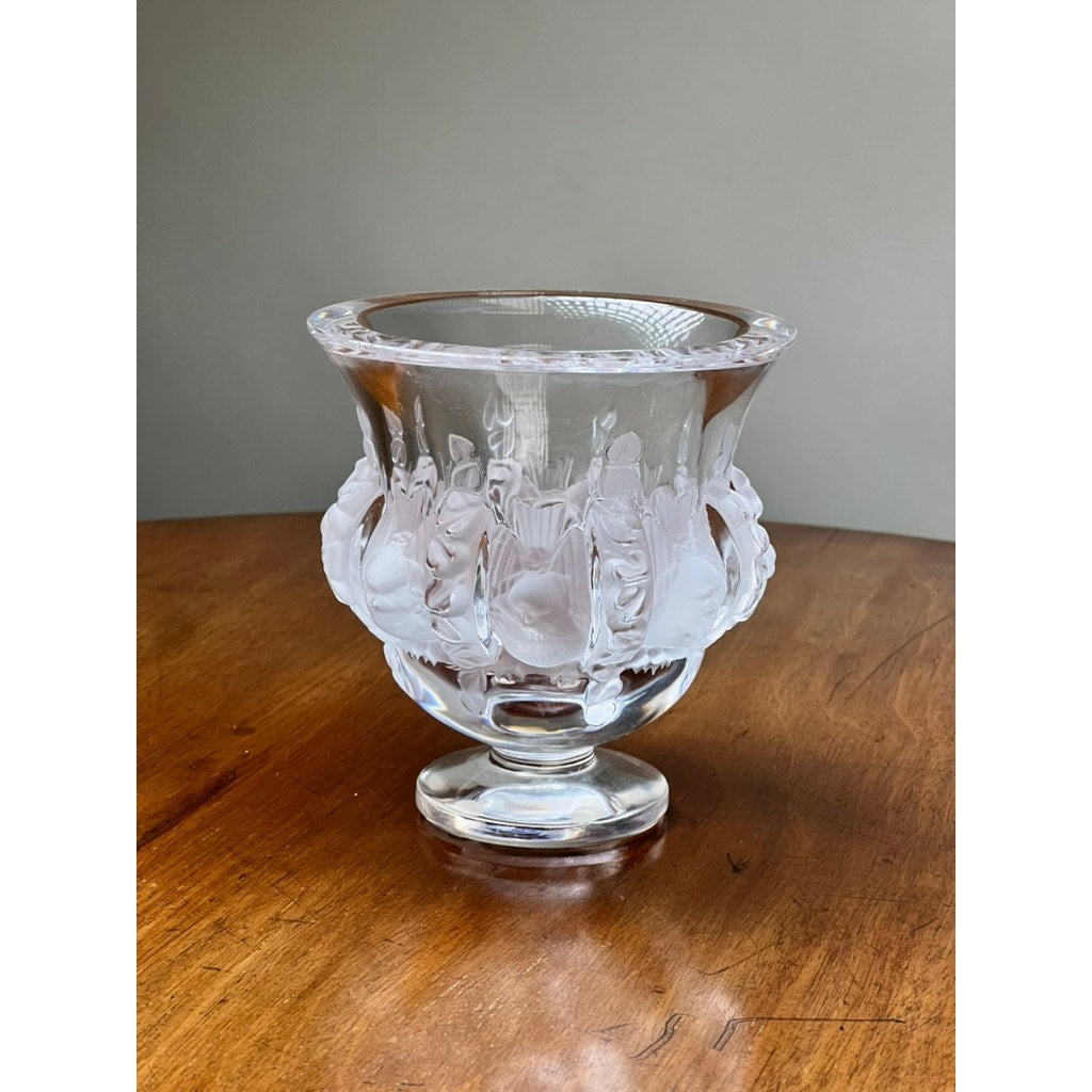 The Vintage Lalique Dampierre Sparrow Footed Vase by Lalique, featuring frosted crystal sparrows and floral motifs, is displayed on a wooden surface against a plain beige background.