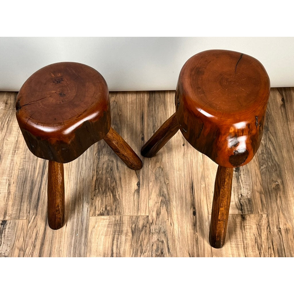 A pair of vintage live edge Wabi Sabi cherry stools, signed by Neil Panyik, display natural wood grain and three polished seats, standing out on a wooden floor against a white wall.