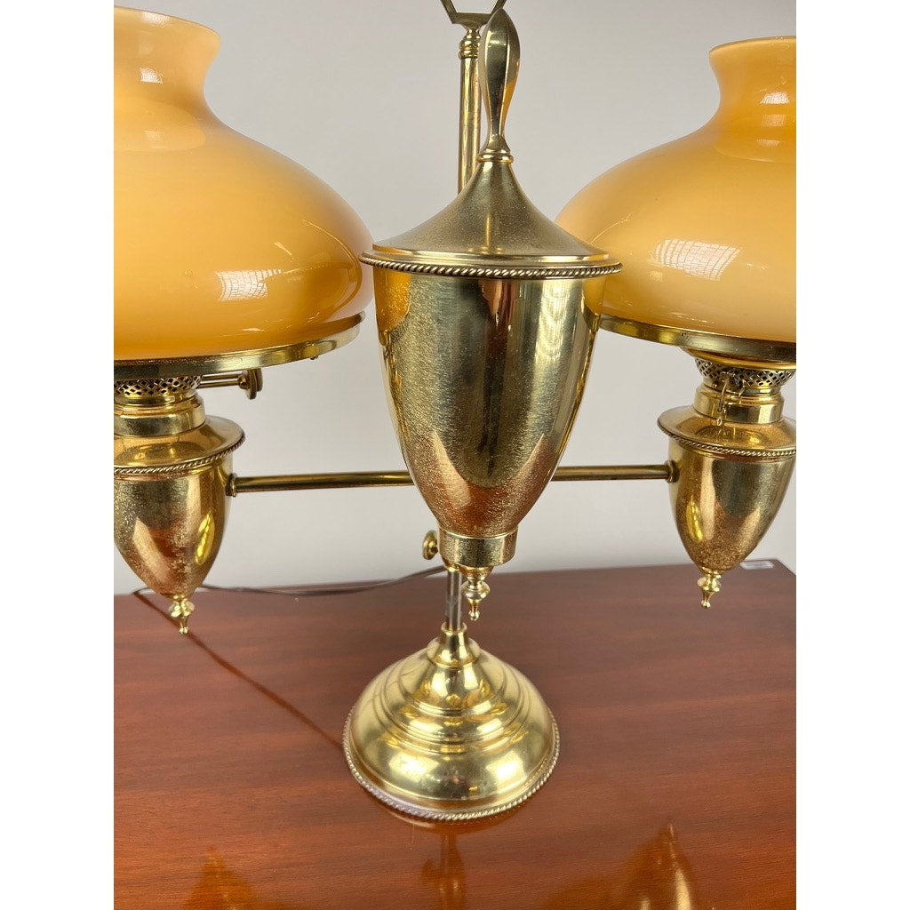 A close-up of an Antique Bradley and Hubbard Brass Double-arm Student Lamp with carmel shades, displayed on a wooden surface against a plain light-colored background.