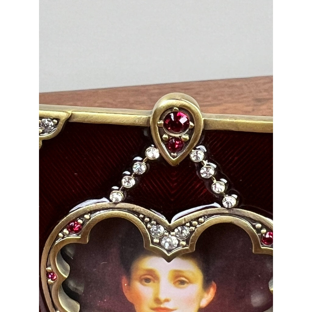 Close-up of the new Jay Strongwater Bejeweled Enamel Double Heart Picture Frame, adorned with red and clear gemstones, featuring part of a painted portrait of a dark-haired woman against a brown and gold background.