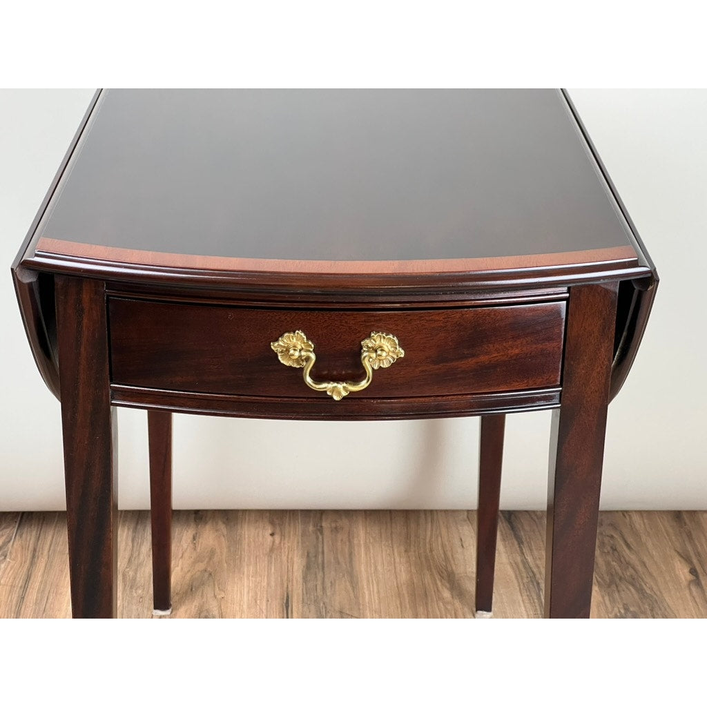 The Vintage Federal Style Edge Banded Mahogany End Table by Stickley features dark wood, a glossy surface, a single drawer with an ornate gold handle, and tapered legs, positioned on a wooden floor against a light wall.