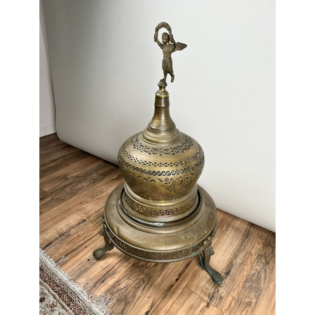 An Antique Venetian Brass Bell Room Heater Brazier, c. 19th Century, with a rounded perforated dome and angel finial stands on an ornate tripod base with curved legs, set on a wooden floor near a patterned rug.