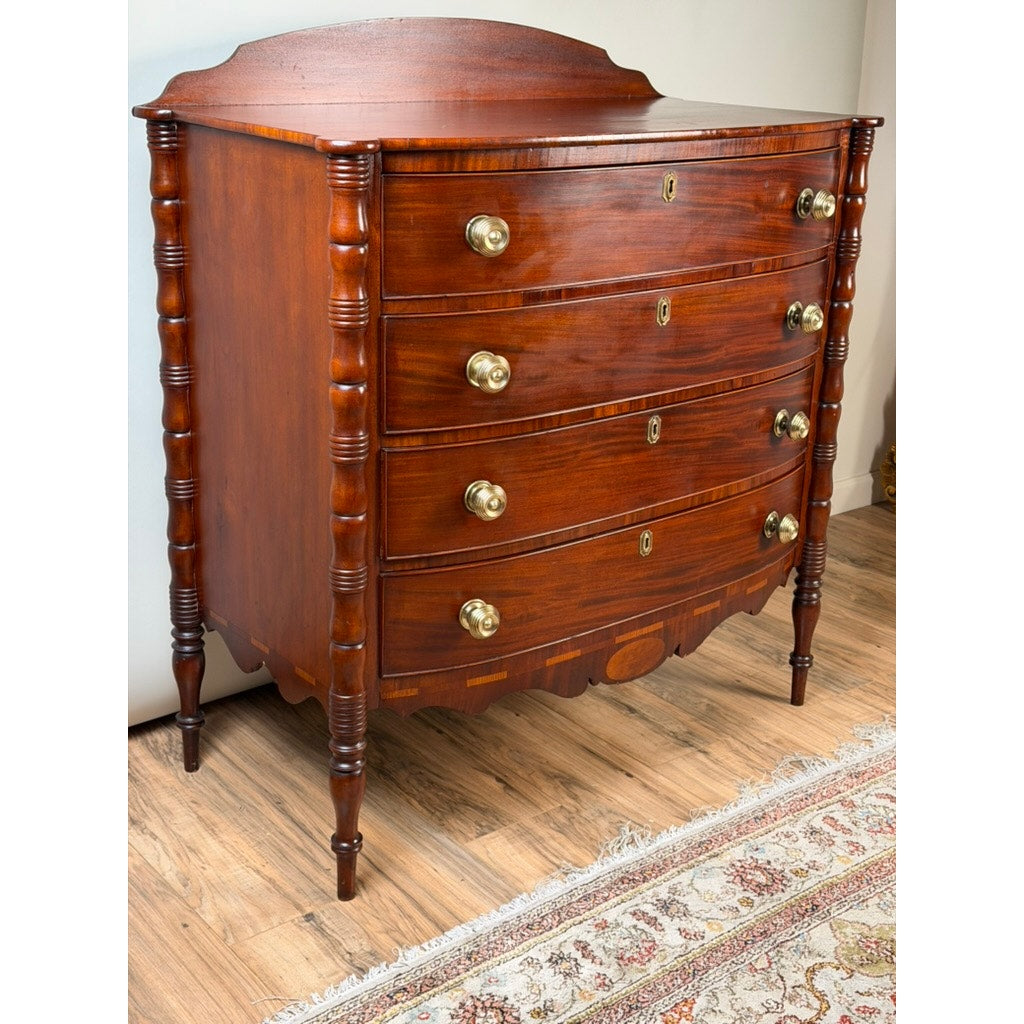 An Early 19th Century New England Federal Mahogany Bow-Front Chest of Drawers with Backsplash, dating from circa 1820–1840, stands gracefully on a hardwood floor next to a patterned rug.