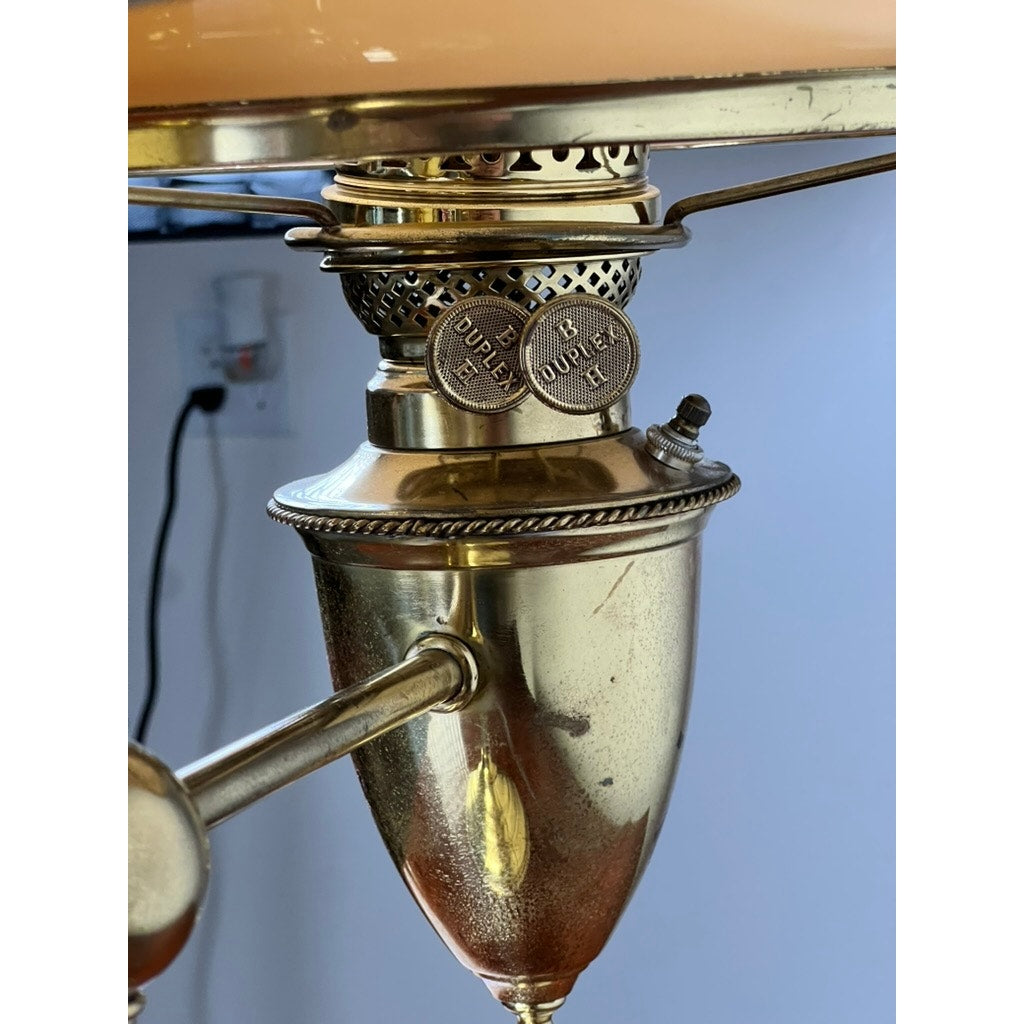 Close-up of an Antique Bradley and Hubbard Brass Double-arm Student Lamp with caramel shades, featuring intricate detailing and two round adjustment knobs, with a blurred power outlet in the background.