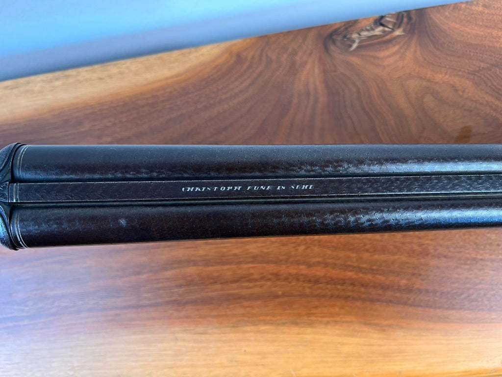 The spine of a dark leather-bound book resting on a wooden surface, with the text ANTIQUE CHRISTOPH FUNKE SUHL GERMAN DAMASCUS DOUBLE BARREL HAMMER SHOTGUN, CARVED STOCK, C. 1880 printed in small white letters along the center, evokes the classic elegance found in antique double barrel shotguns offered by Pearl City Antiques.