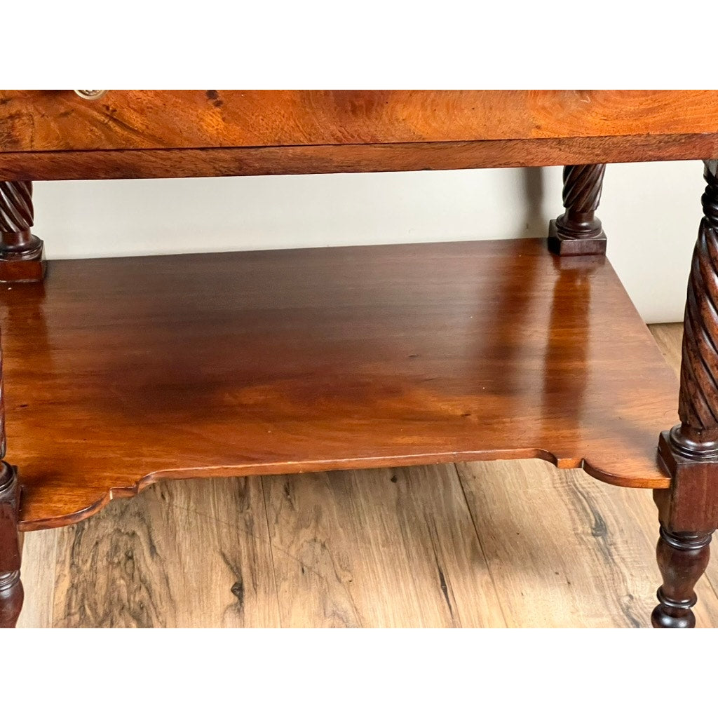 An Antique Federal Mahogany Two Drawer Work Stand with lions head pulls, c. 1820, featuring decorative carved edges and four spiral-carved legs, stands on a wooden floor against a plain wall.