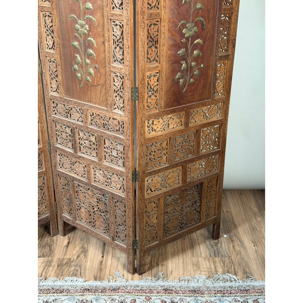 An Early 20th Century Anglo-Indian Carved Teak Four-Panel Screen with Applied Brass Floral Decoration by Pearl City Antiques stands elegantly on a wooden floor beside a patterned rug.