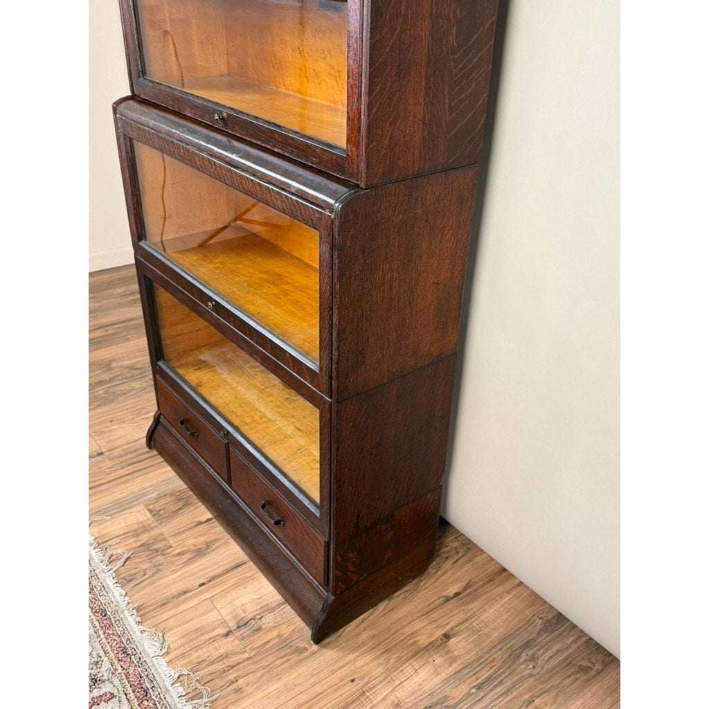 An Antique Hale System Quarter-Sawn Oak Five-Stack Barrister Bookcase by Camden Cabinet Company, dating to circa 1910, stands on a wooden floor beside a light-colored wall and a patterned rug. Featuring glass-fronted shelves and two lower drawers, its classic design and sturdy craftsmanship capture the timeless appeal of an authentic antique barrister bookcase.