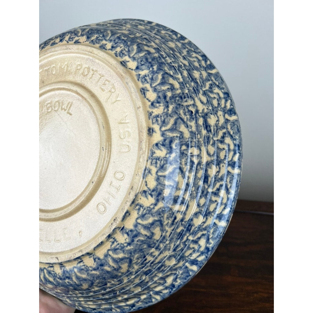 A hand holds an upside-down Robinson Ransbottom Roseville Blue Spongeware Pasta Bowl with a blue and cream marbled pattern. The unglazed base is stamped “ROSEVILLE, O. OHIO, USA” and “ROBINSON RANSBOTTOM.”.